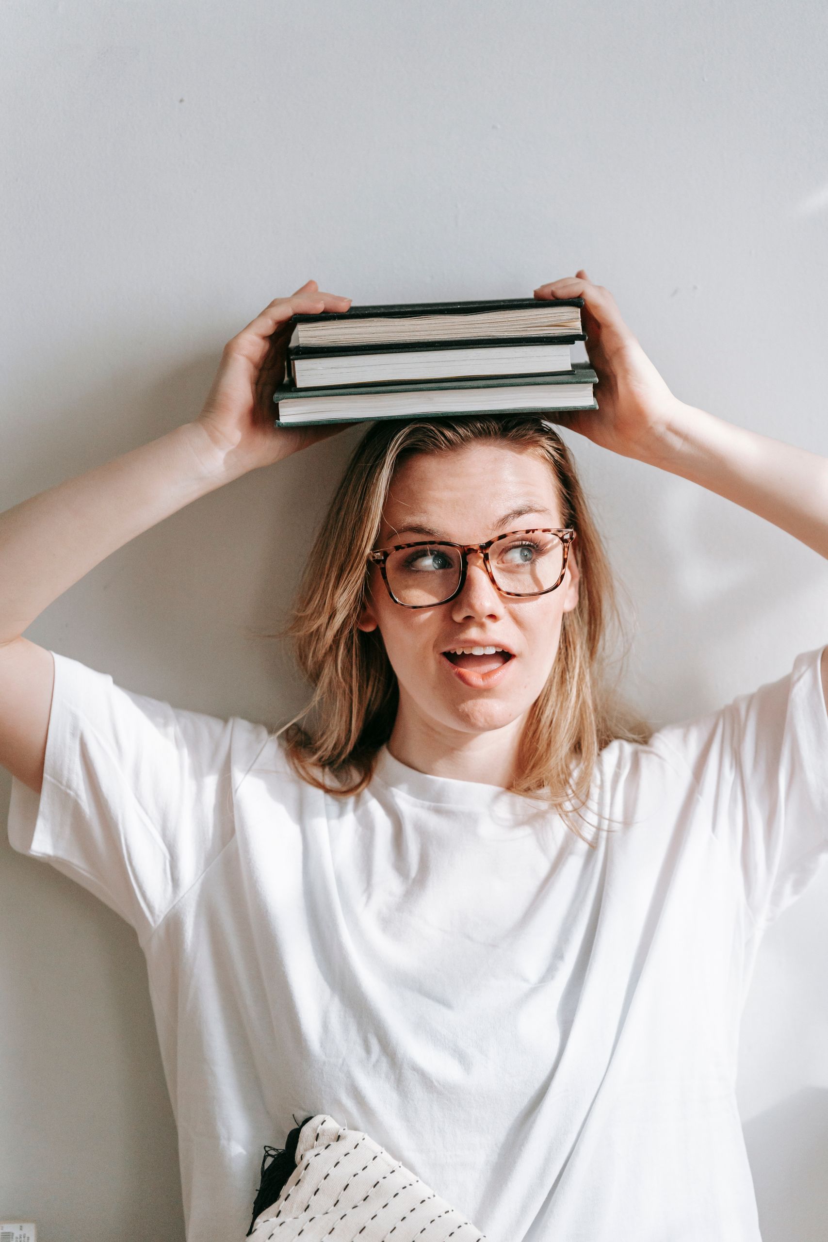 Cheerful young female in casual t shirt looking away while sitting on white background with collection of books on head