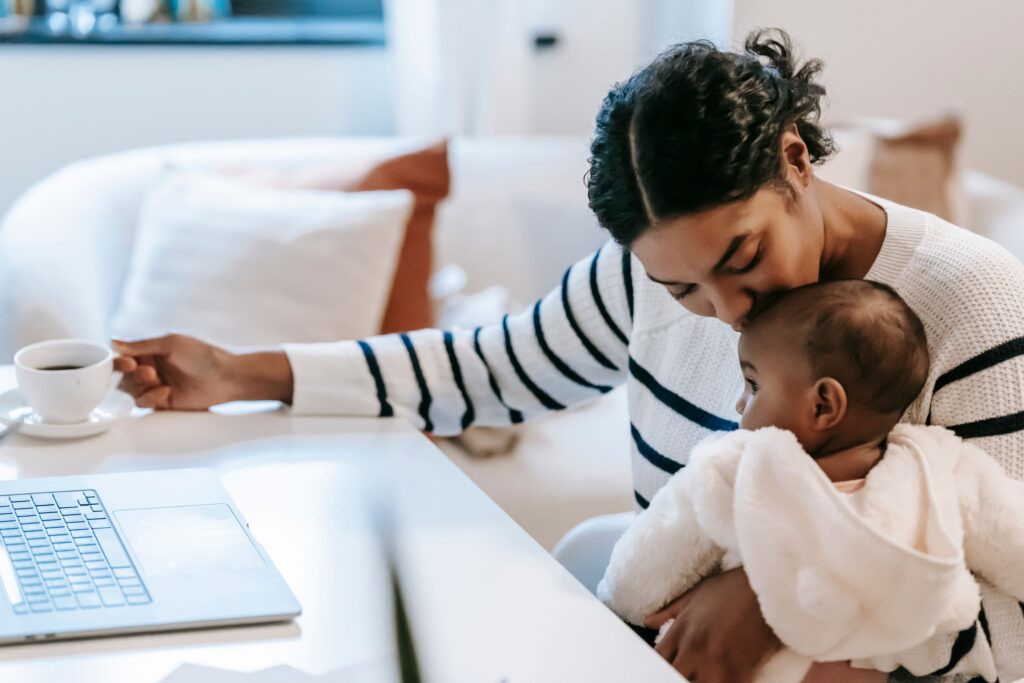 Mother hugging and kissing in head toddler on laps in casual clothes near table with laptop and mug with tea while sitting in chair at light room near couch